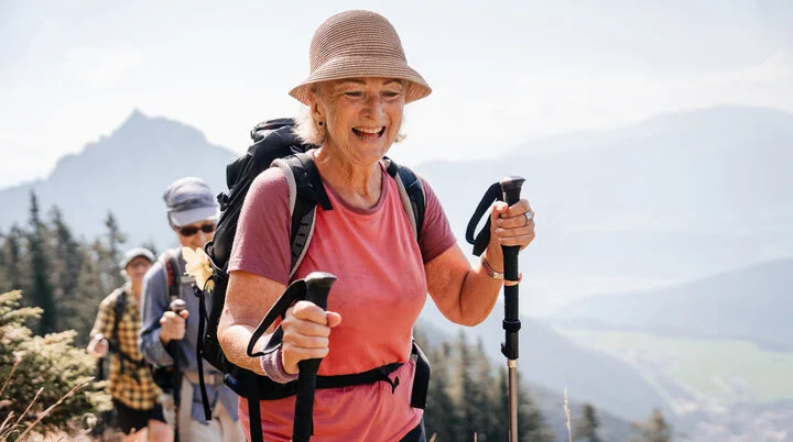 Eine ältere Frau mit Stöcken und Sonnenhut führt glücklich eine Wandergruppe an. | © DAV / Marisa Koch